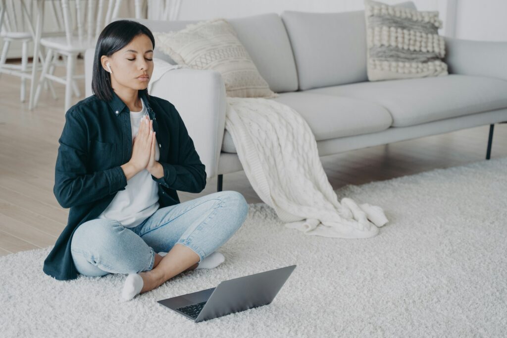 Calm woman meditates to relieve stress, practicing yoga at laptop in lotus asana on floor at home