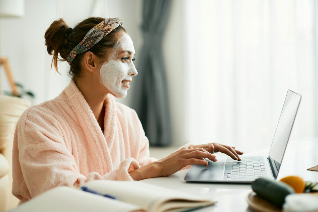 Young woman in bathrobe surfing the net on laptop while wearing cosmetic face mask.