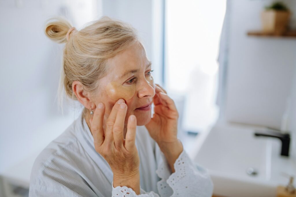 Beautiful senior woman in bathrobe, applying eye patches for puffiness while looking in the mirror